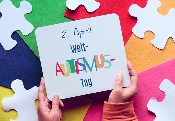 H&auml;nde halten Seite mit dem Text 2. April Welt-Autismus-Tag. Im Hintergrund sind Puzzle-Element auf geschichtetem, buntem Filzhintergrund.